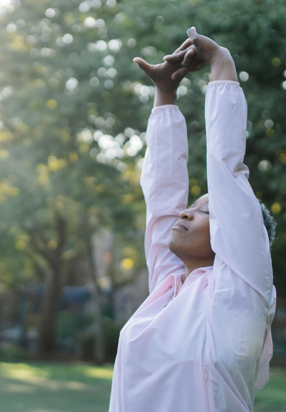 Woman stretching A Black, middle aged woman stretches her arms up overhead with her fingers clasped together and palms facing up. Her eyes are closed and her face is tilted up towards the sun. She appears to be standing in front of a forest of trees.